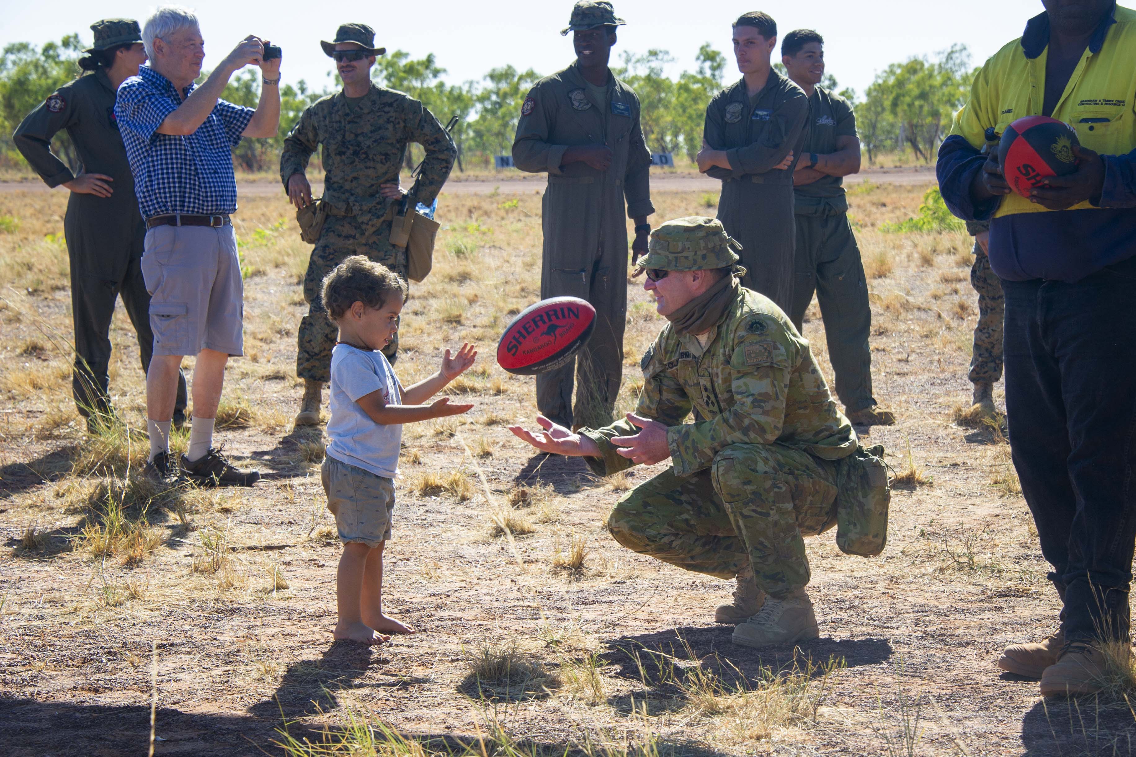 An Australian soldier in camoflague gear knees on the dirt and throws preschooler a Sherrin AFL football.