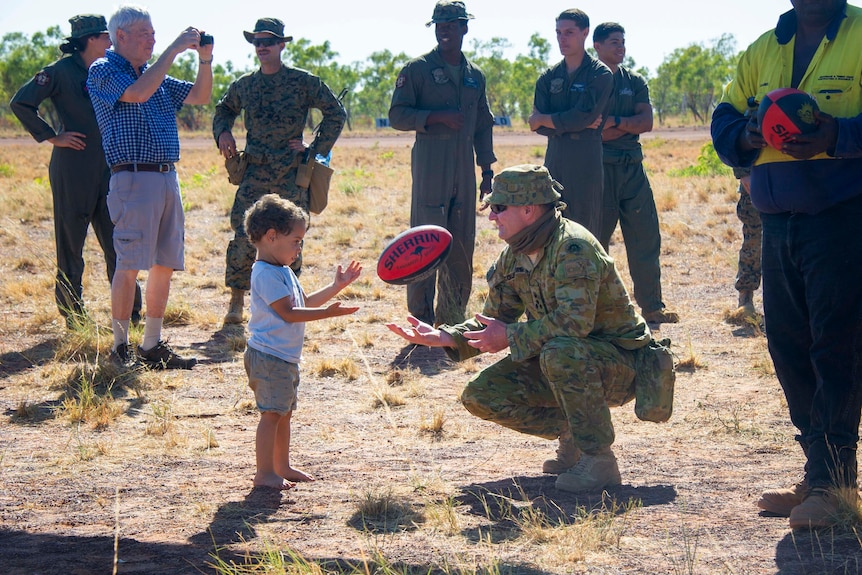 An Australian soldier in camoflague gear knees on the dirt and throws preschooler a Sherrin AFL football.