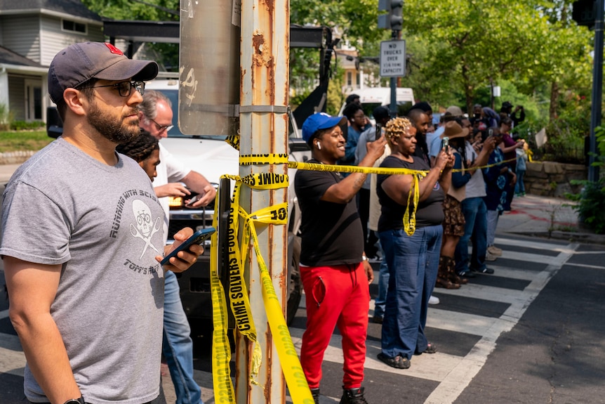 A row of people watch, some pointing mobile phone cameras, from behind a police tape