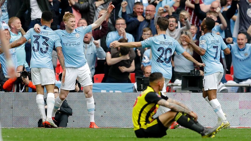 Kevin de Bruyne holds his hand in the air and smiles as his teammates run towards him