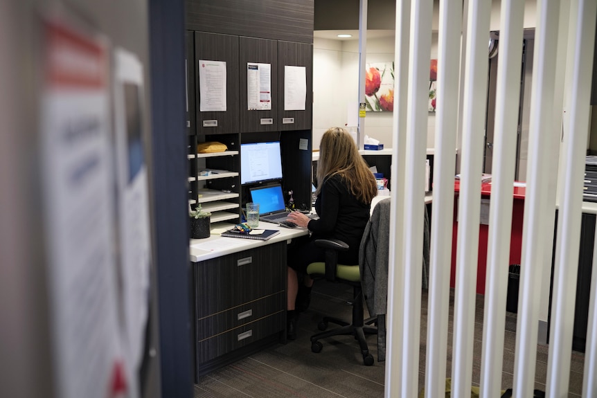 The blonde haired receptionist has her back to the camera and is sitting at her desk working on her computer. 