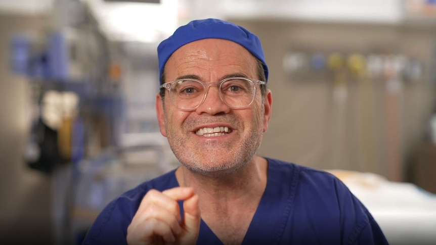 Dr Lanzer, dressed in surgeon's scrubs, gestures and smiles at the camera, sitting in what appears to be a surgery room.