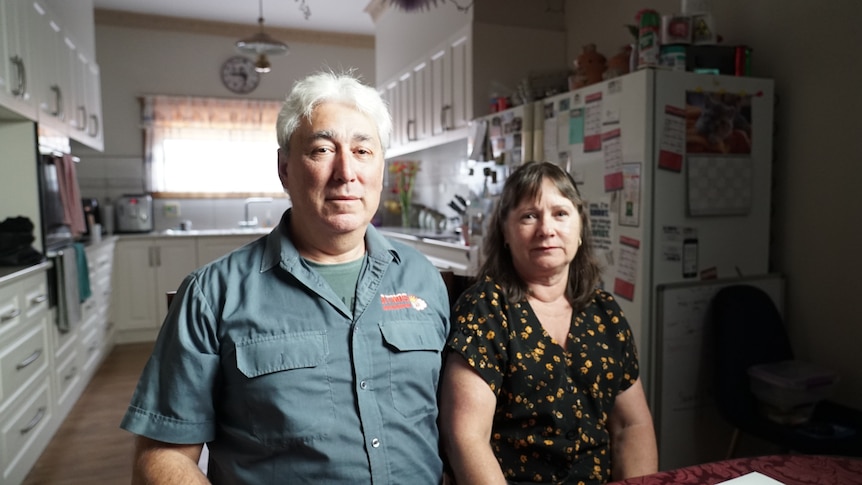 Tony and Leanne Nesci, a middle-aged couple, are sitting at a table in a ktichen and looking seriously into the camera.