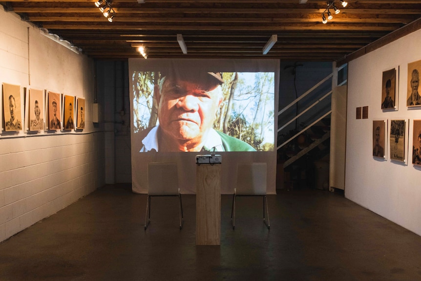 Portraits on a gallery wall and large projector screen in centre with a man's face