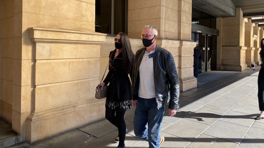 A woman in a black coat, dress and tights walks beside a man in a leather jacket outside a court building