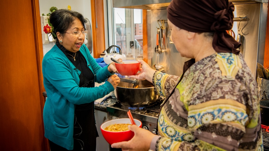 Thit smiling as she ladles some laksa soup into a bright red bowl, for a woman wearing a head covering.
