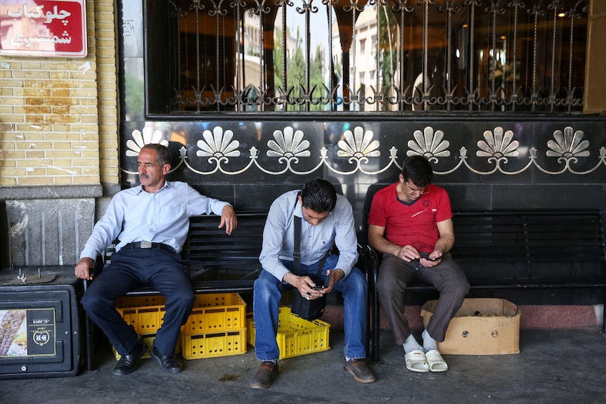 Three men sit on a bench outside a building, two of them looking down at smartphones.