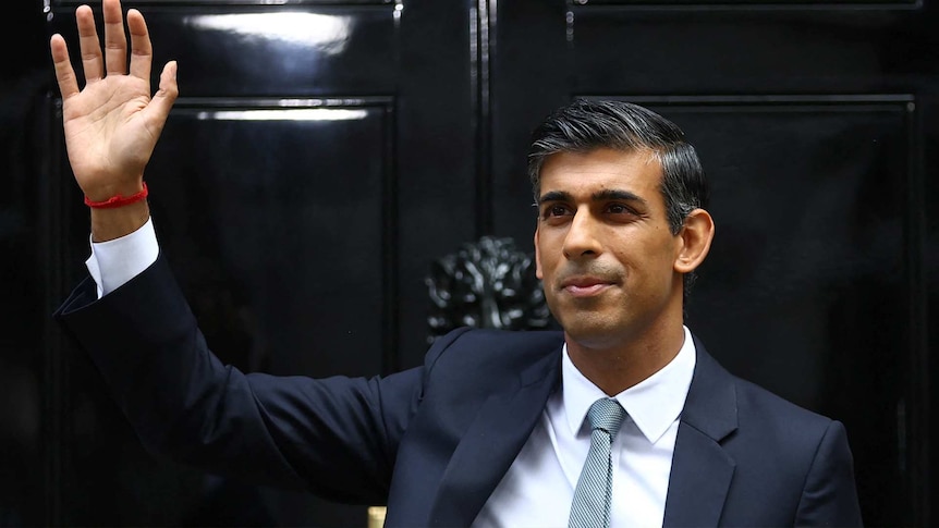 Rishi Sunak wearing a suit and smiling waves from the door of 10 Downing Street.