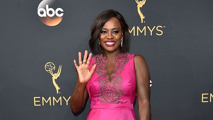 Viola Davis waves to the camera on the red carpet wearing a magenta dress with a lace neck detail