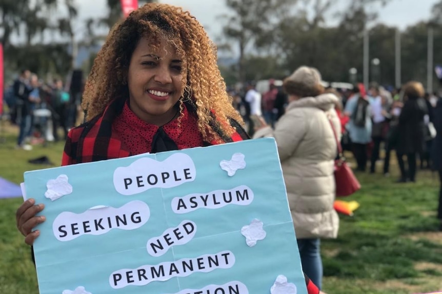 A woman holding a sign that reads 'people in detention need permanent accommodation'.