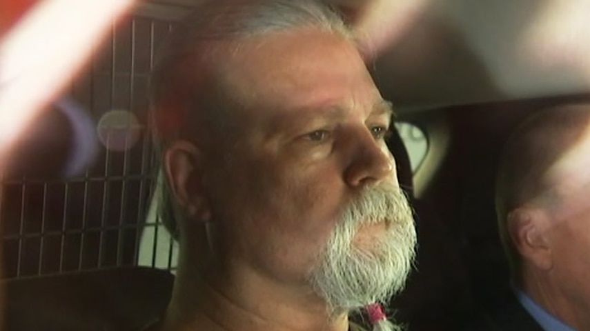 A bearded man in the back seat of a police car, a detective beside him