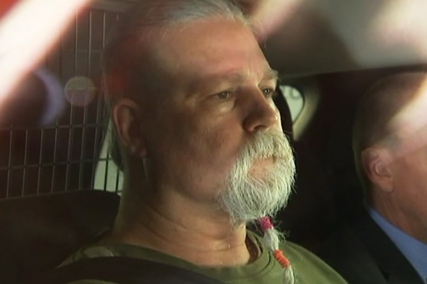 A bearded man in the back seat of a police car, a detective beside him.