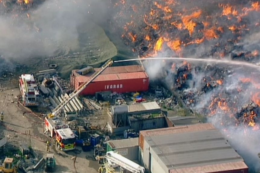 An aerial photo of a huge fire burning in an industrial area, with fire trucks spraying water on it.
