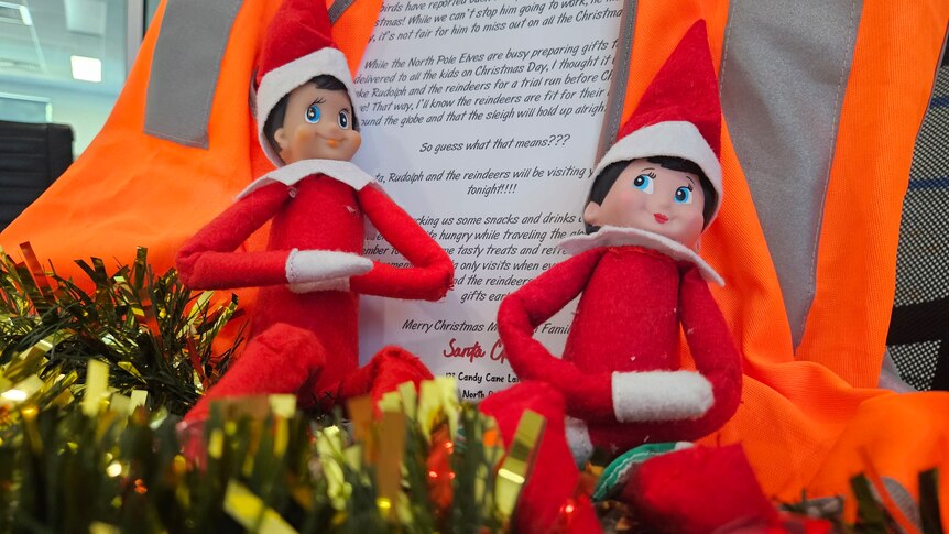 Two elves sit on tinsel in front of a hi-vis vest and a note that says Santa will be coming early