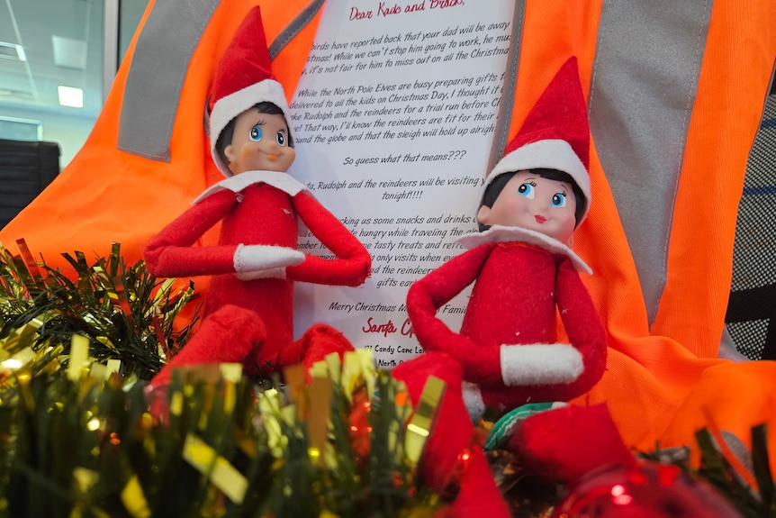 Two elves sit on tinsel in front of a hi-vis vest and a note that says Santa will be coming early