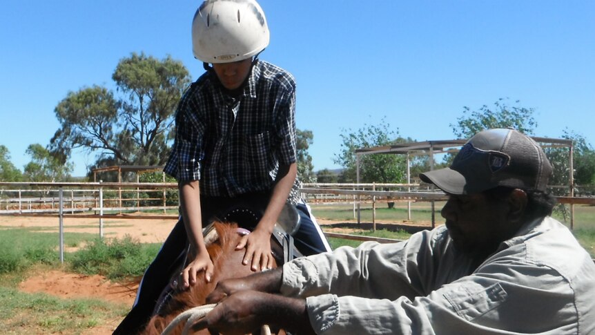 A young Aboriginal boy on horseback is being given instructions from a teacher standing next to the horse.