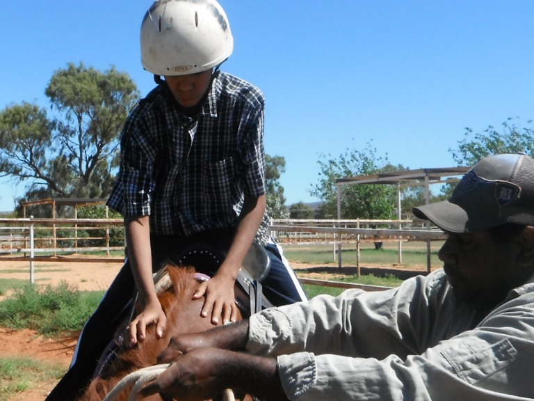 A young Aboriginal boy on horseback is being given instructions from a teacher standing next to the horse.