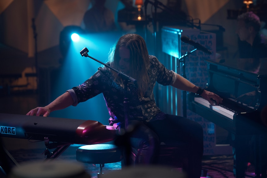 A man backlit by stage lights plays one piano with each hand.