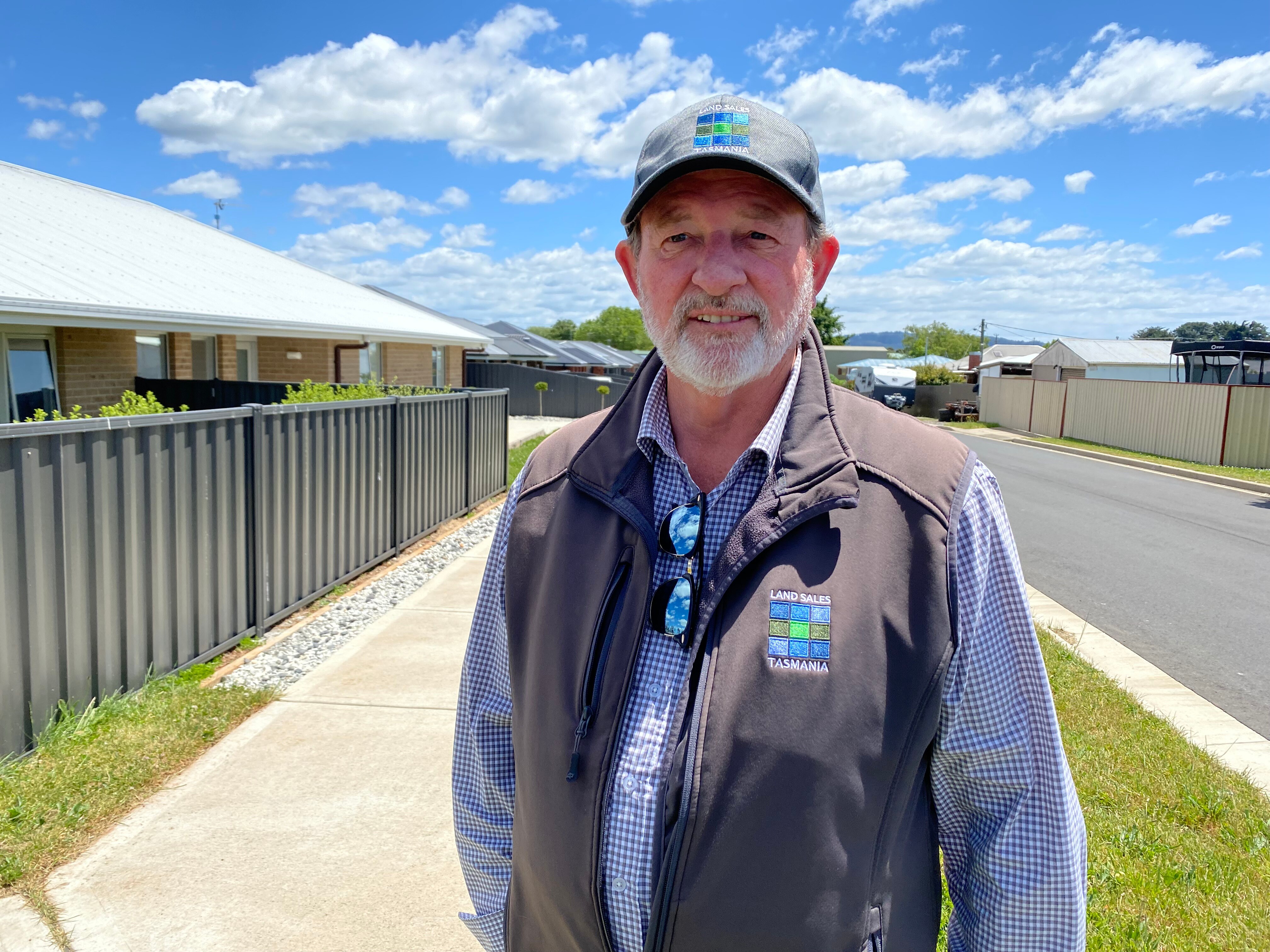 A middle-aged man wearing a cap standing on a sidewalk in front of newly built houses on a sunny day.