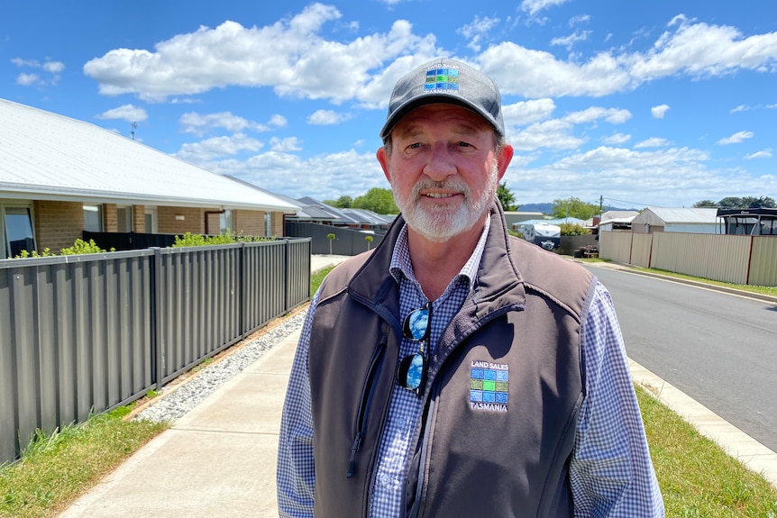 A middle aged man wearing cap standing on a sidewalk in front of newly built houses on a sunny day.