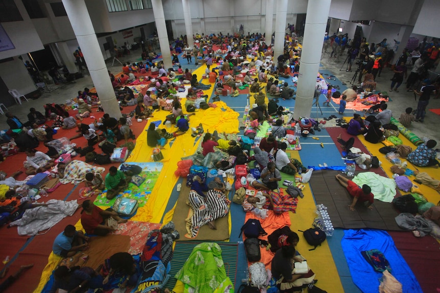 A photo from high up shows an overview of people sitting on the ground with blankets in a hall.