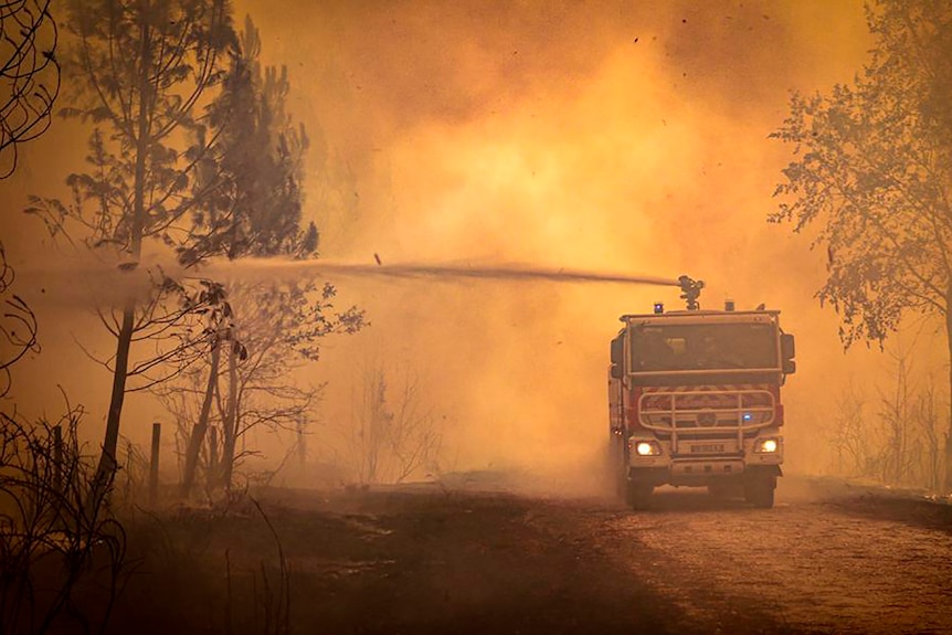 Un camion de pompiers pulvérise de l'eau dans un feu de forêt, alors que la fumée l'entoure et la route.