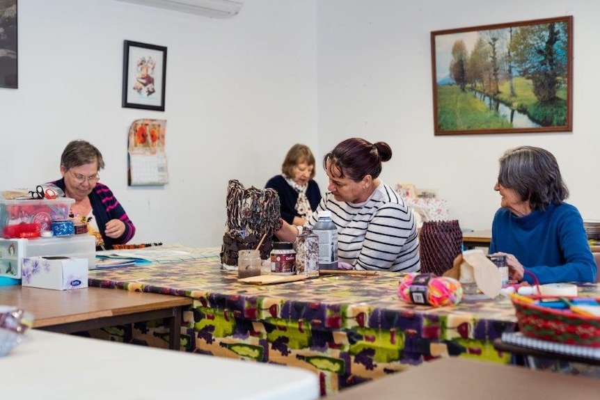 Four women seated at a table in the room of a house working on crafts.