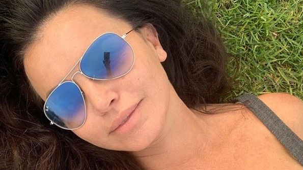 Suzi Taylor lays on grass in a close head shot, she is taking a selfie and wears aviator glasses.