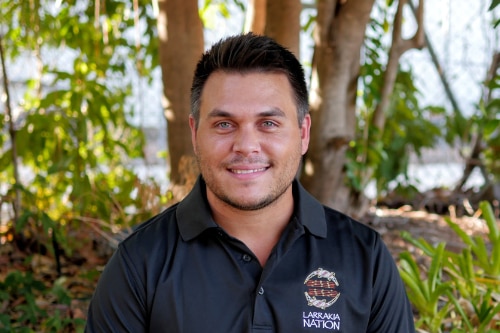 A young man in a black polo shirt sits with plants behind him. He is smiling at the camera