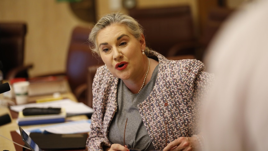 A blonde woman wearing a maroon patterned jacket jacket sitting at a table mid-sentence