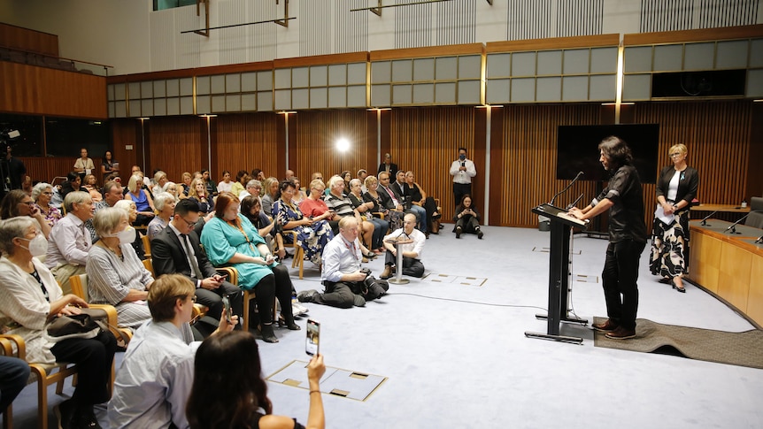 Boochani stands at a lectern looking out to a seated crowd.