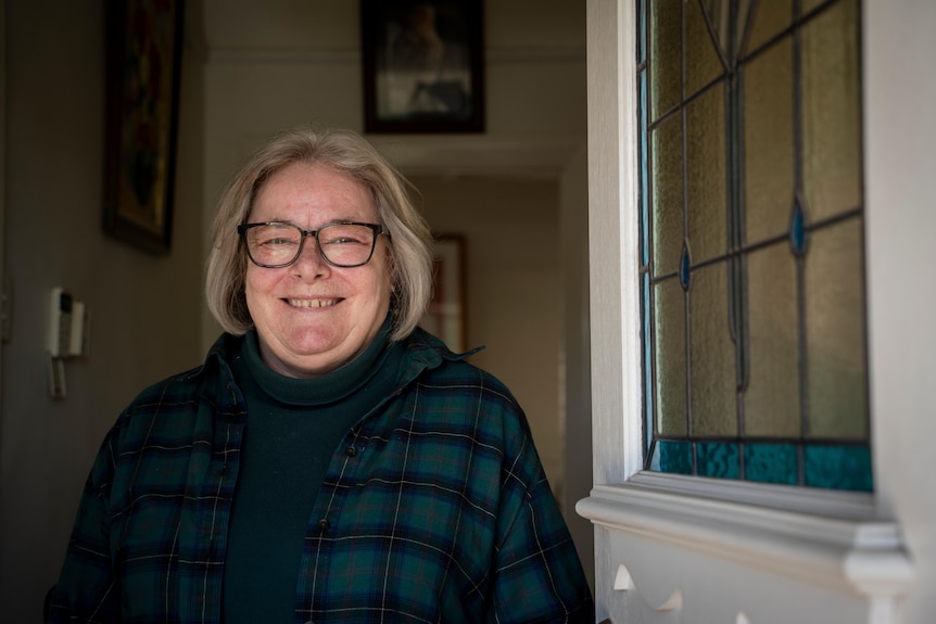 Christine Thirkell wearing black framed glasses and a checkered jacket standing next to her front door smiling. 