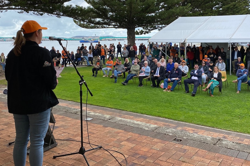 A photo taken from a stage behind a speaker, showing a large crowd of people seated in front of her in a park setting.