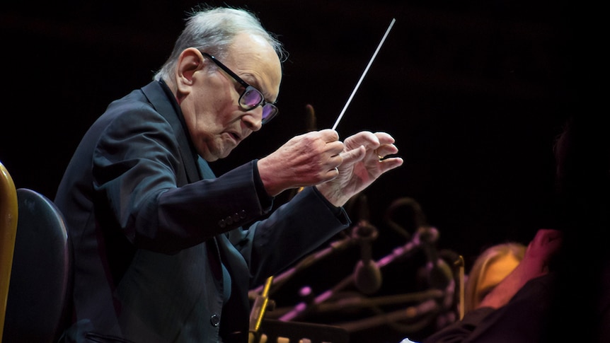 Ennio Morricone conducts on stage. He wears glasses with dark frames and holds a baton in the air.