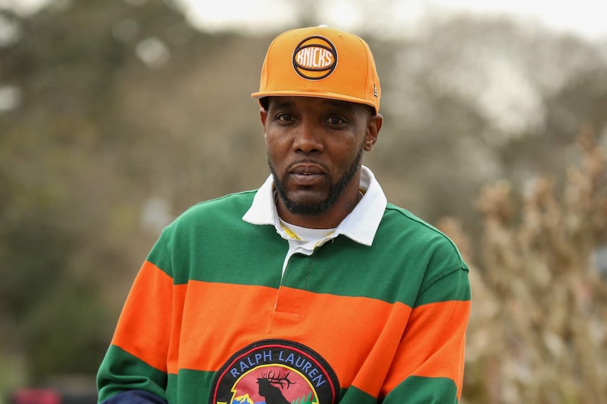 A man in a Knicks cap and Ralph Lauren polo leaning on a fence looking solemn