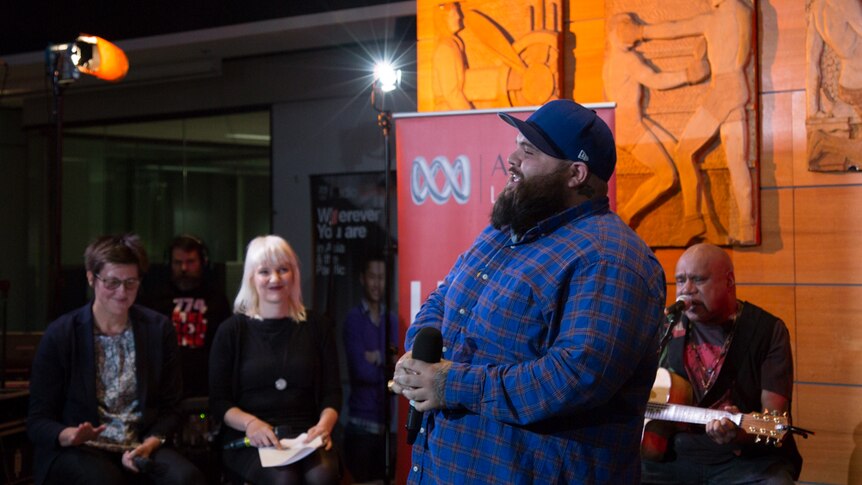 Two women watch a standing man and a sitting man perform. Man in headphones in background.