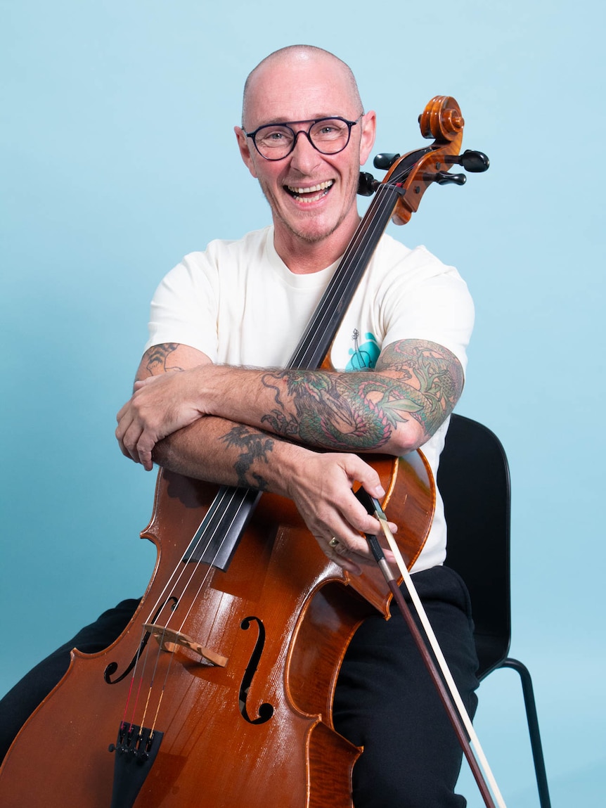 Ed Le Brocq sits smiling at his cello in a white tshirt and black jeans.
