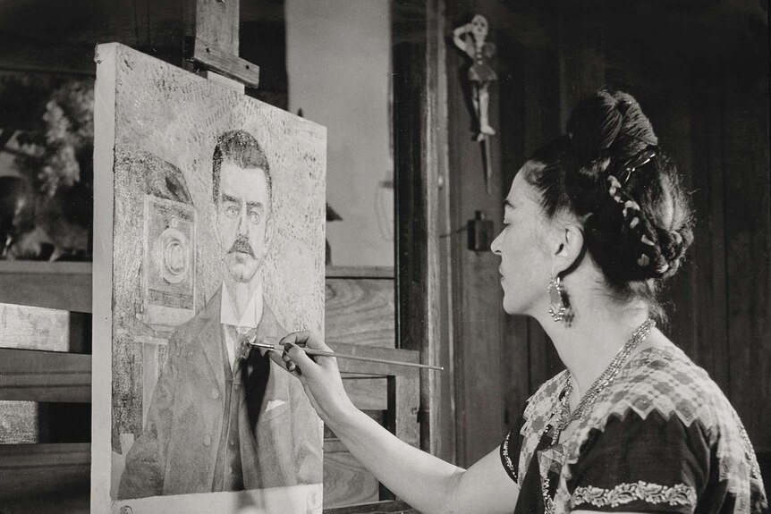 Frida Kahlo works on a portait of her father, Guillermo. She's seen wearing her traditional dress and hair tied up in braids.