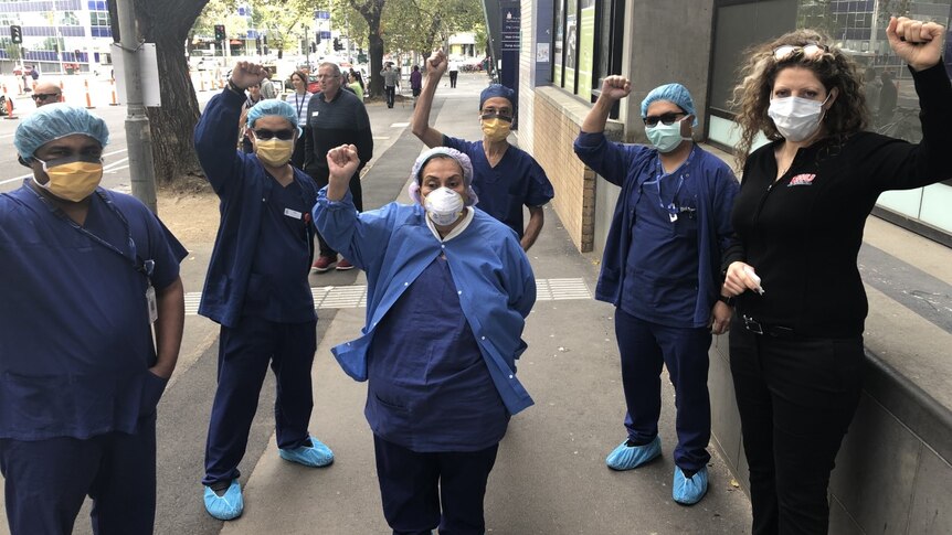 A group of health care workers in blue hold their fists up wearing PPE on the street in a grey day.