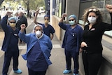 A group of health care workers in blue hold their fists up wearing PPE on the street in a grey day.
