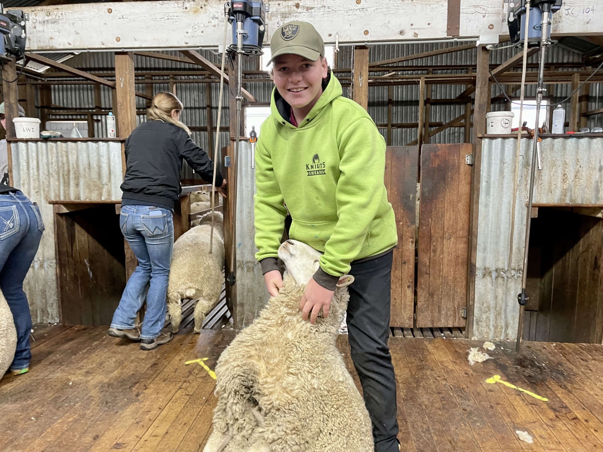 A teenage boy in a yellow/green jacket holding a sheep in a sheairng shed, Crookwell NSW, June 2023