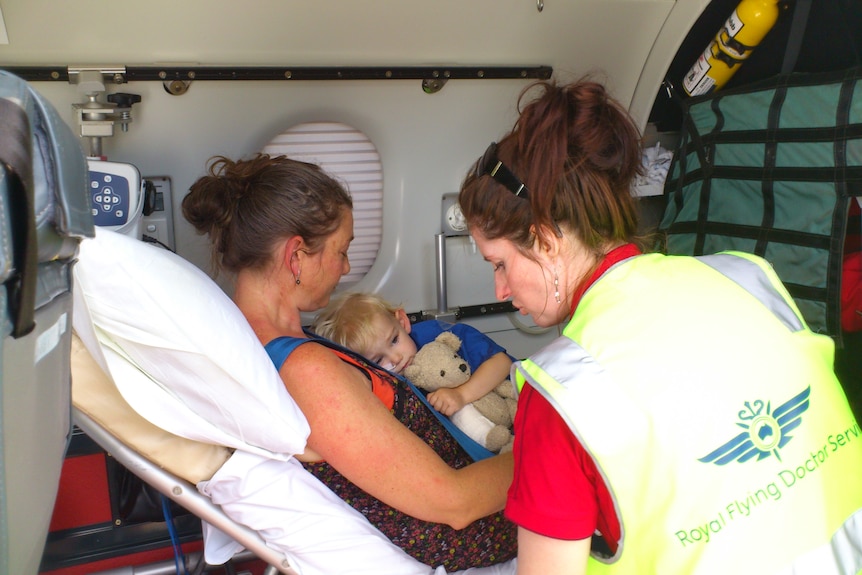 Woman in van lies on hospital bed with young boy grasping teddy bear on her chest, woman in yellow high-vis vest in front.