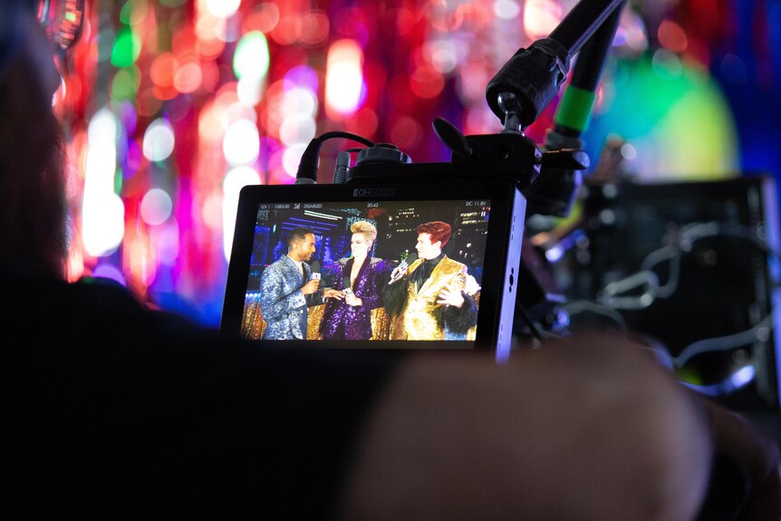 A camera monitor shows three people dressed in glamourous clothing speaking with a microphone
