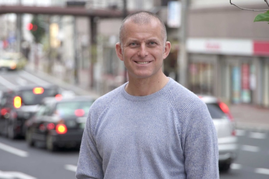 A man in a grey shirt with a blurred busy street in the foreground.