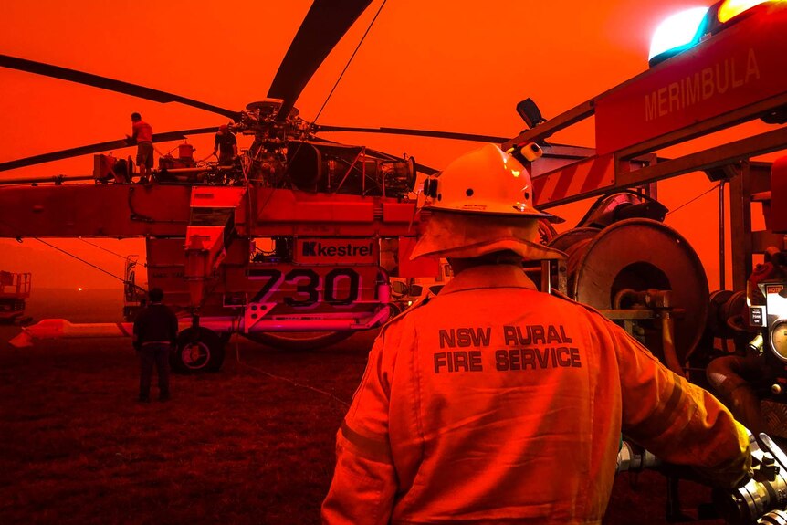 Rear of person in Rural Fire Service uniform watching as large helicopter is serviced, entire scene is tinged red by bushfires.