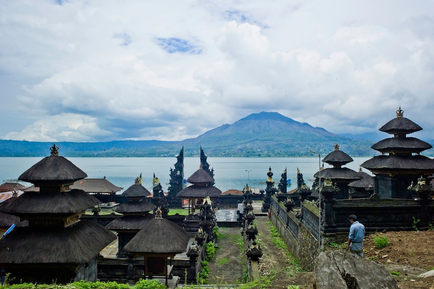A man in a blue shirt walks through a series of Balinese buildings, he is facing a lake and a volcano.