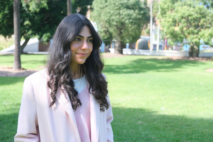 A woman with long dark hair and pink blazer looking off camera while standing in a park.