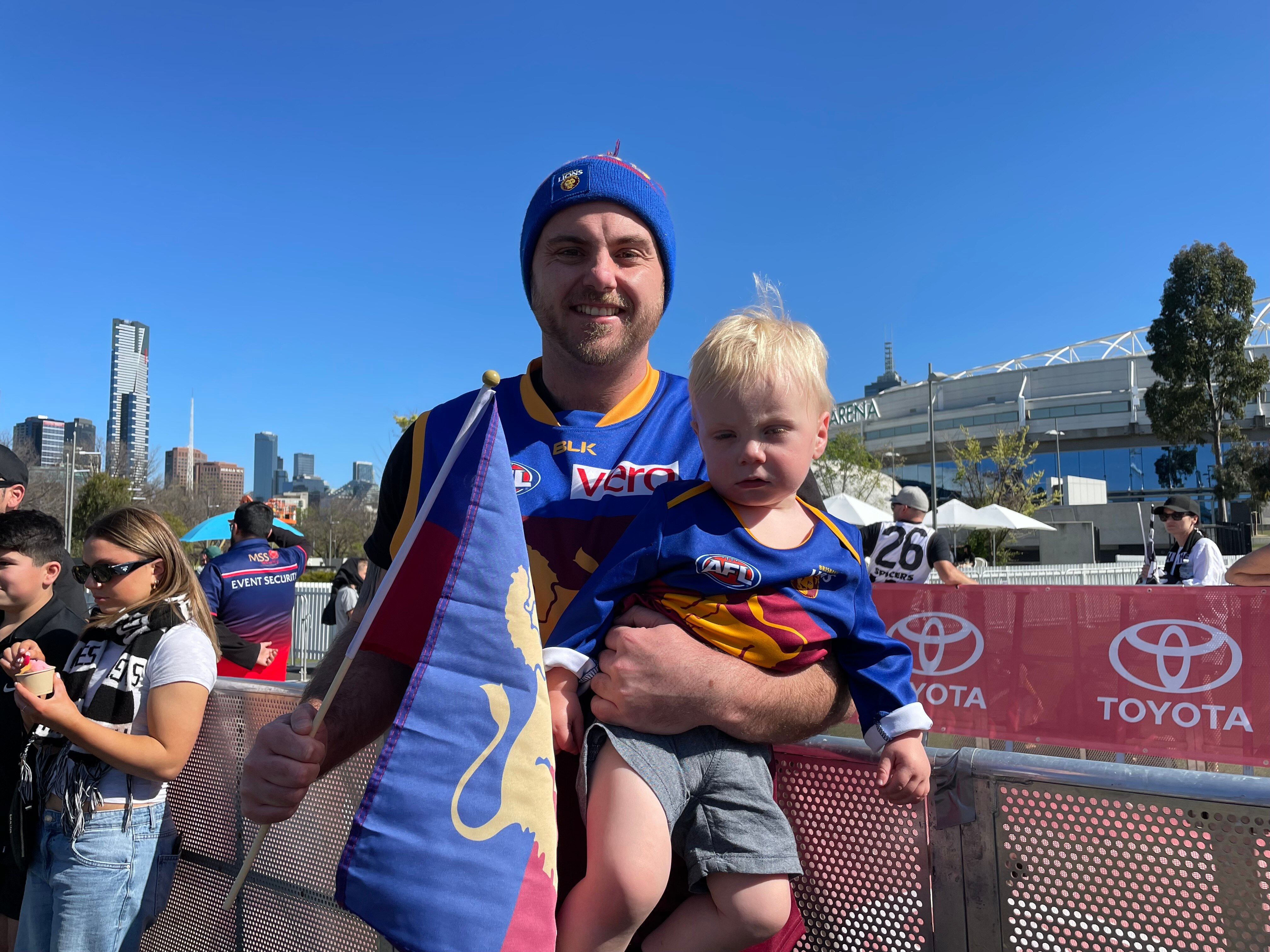 A man wearing Brisbane Lions colours holds up a toddler.
