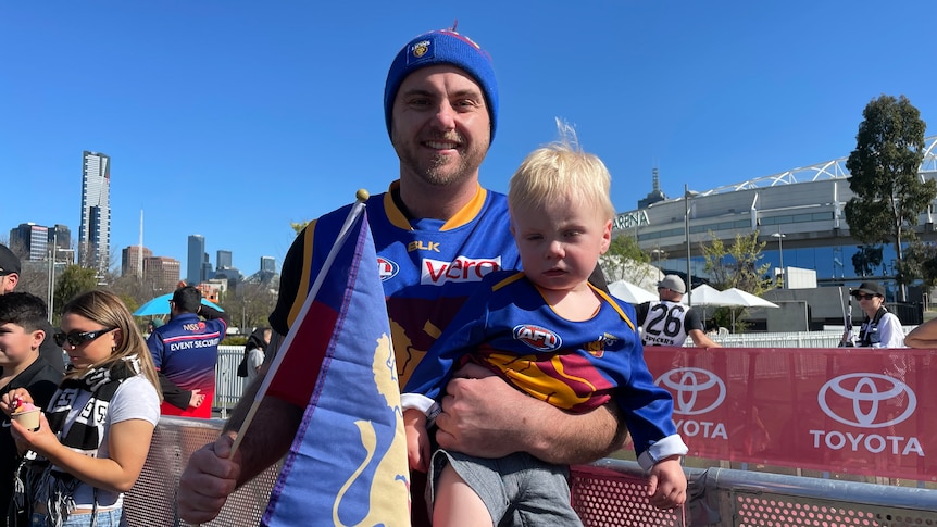 A man wearing Brisbane Lions colours holds up a toddler.