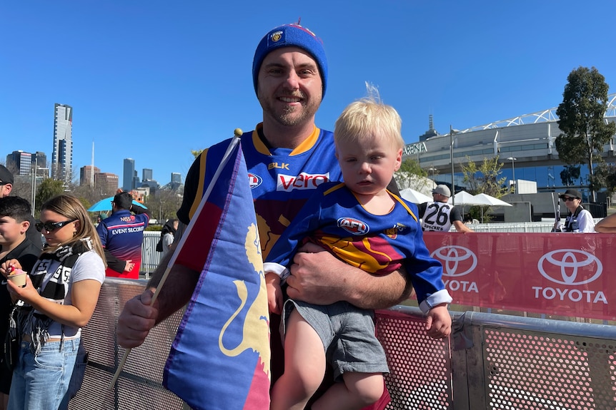 A man wearing Brisbane Lions colours holds up a toddler.